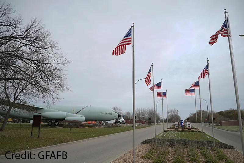 A row of American flags on tall poles lines a road at Grand Forks Air Force Base, with a U.S. Air Force aircraft on static display in the grass to the left and bare trees under an overcast sky.