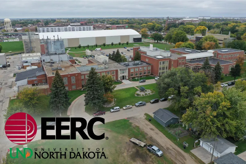 An aerial drone view of the University of North Dakota's Energy & Environmental Research Center (EERC) campus, showing a complex of red-brick research buildings, industrial structures, and a large white warehouse, with parking lots, green lawns, and a water tower visible in the background, overlaid with the EERC and University of North Dakota logos in the lower left.