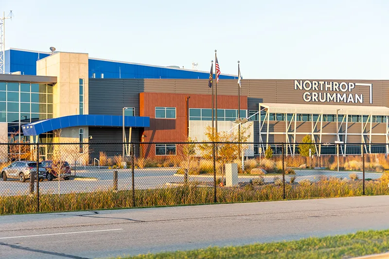 The exterior of the Northrop Grumman facility at Grand Sky technology park, featuring a modern building with blue, brown, and rust-colored architectural panels and large windows, the Northrop Grumman logo on the right side, American and state flags flying out front, and a chain-link fence in the foreground at golden hour.