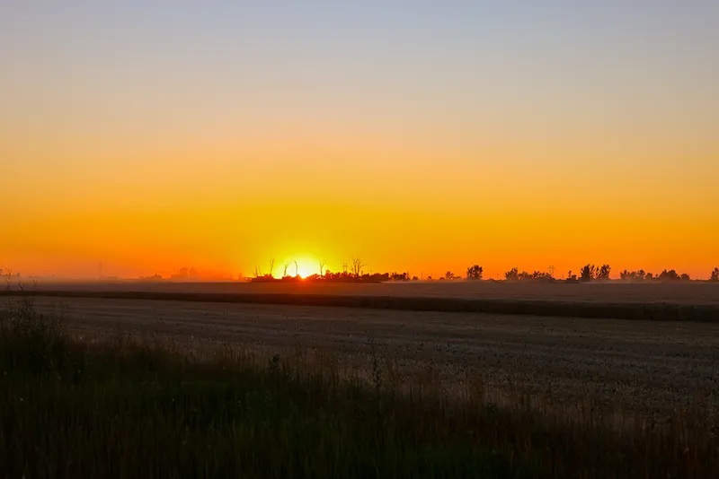 A vivid orange and yellow sunset over flat, harvested agricultural fields in the Red River Valley, with silhouetted trees along the horizon.