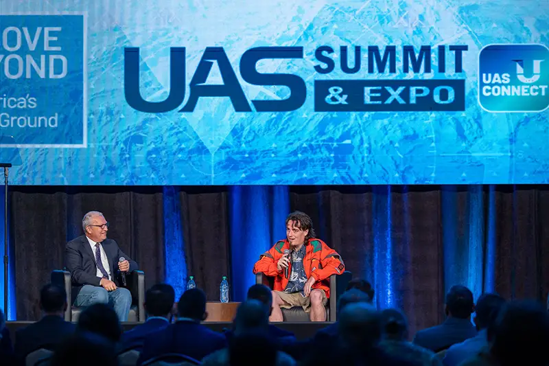 Two men in conversation on stage at the UAS Summit & Expo, with a large blue screen behind them and an audience seated below.