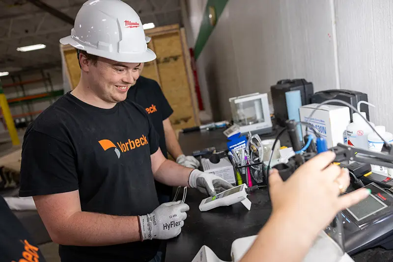 A smiling young man wearing a white hard hat, HyFlex gloves, and a black Vorbeck t-shirt works at an industrial workbench, handling a small device alongside a colleague.
