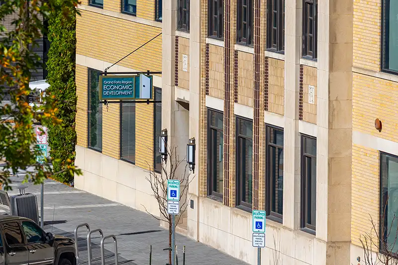 Street-level view of a yellow brick building in downtown Grand Forks displaying a hanging sign for the Grand Forks Region Economic Development office.