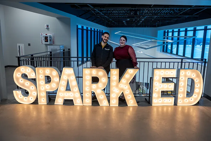 A man and a woman stand smiling behind large illuminated marquee letters spelling "SPARKED" at the EDND Spring Conference 2026 in Bismarck, with bright white bulbs lighting up each letter and a modern industrial interior with metal railings and blue lighting in the background.