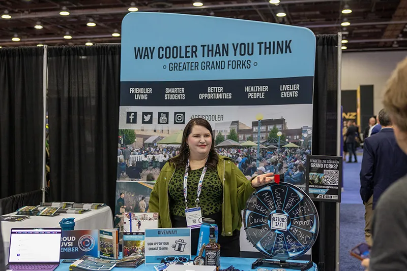 A young woman with long dark hair, wearing a dark green jacket over a patterned top, stands at the Greater Grand Forks "Way Cooler Than You Think" trade show booth at AUVSI Xponential in Detroit, holding a spinning prize wheel beside a table of promotional materials and a giveaway sign.