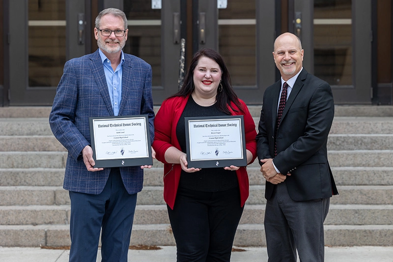 Three people stand smiling on the steps of a building, with two of them holding framed National Technical Honor Society certificates; a man with glasses and a graying beard in a blue plaid blazer on the left, a woman with long dark hair in a red blazer over a black top in the center, and a man in a dark suit with a red tie on the right.