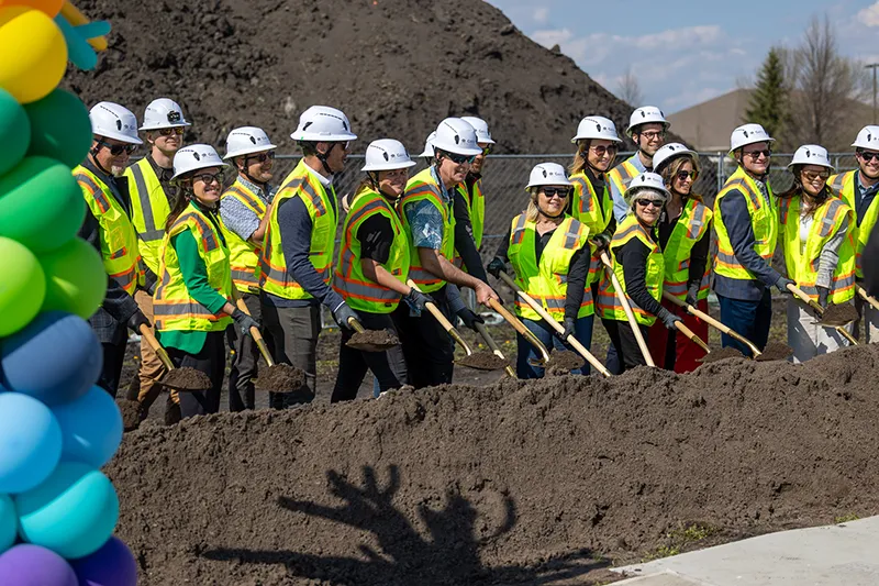A long row of community leaders and stakeholders wearing white hard hats and bright yellow safety vests stand together holding shovels in piles of dirt during the Children's Museum groundbreaking ceremony, with colorful rainbow balloons on the left side and a mound of soil in the background.
