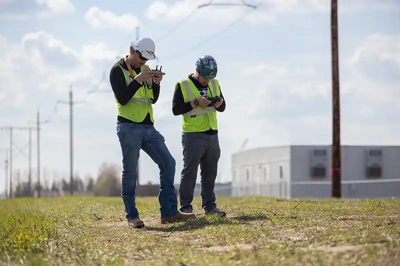 Two UAS drone pilots stand in a grassy field on a partly cloudy day, both wearing bright yellow-green high-visibility safety vests and operating handheld drone controllers; one wears a white hard hat and looks down at his controller while the other wears a backwards baseball cap and concentrates on his screen, with power line poles, a utility building, and a wooden post visible in the background.