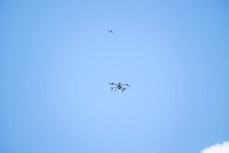 Two drones flying against a clear blue sky, with a larger multi-rotor UAS in the foreground and a smaller drone visible higher in the background.