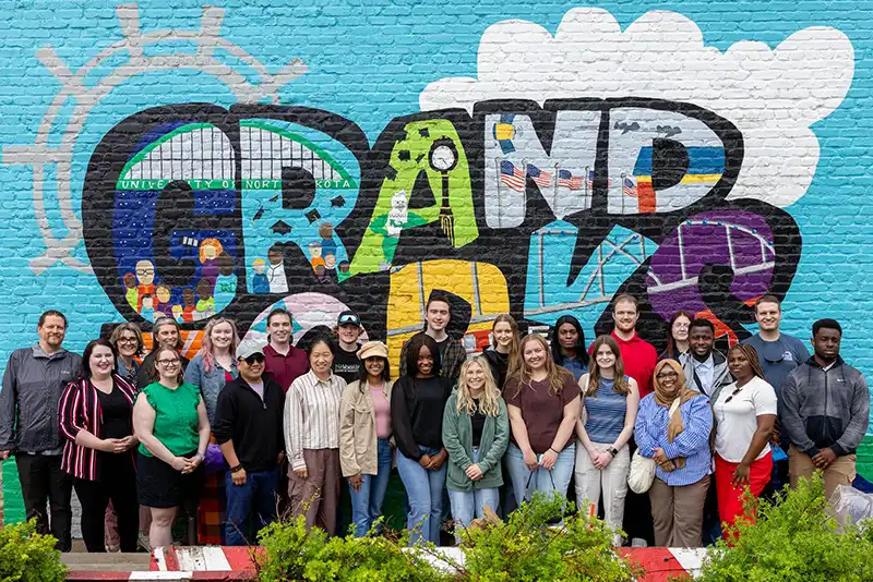 A large group of InternGF summer cohort participants poses and smiles in front of a colorful "Grand Forks" mural painted on a brick wall.