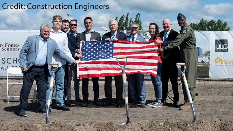 A group of community and military leaders pose with ceremonial shovels and an American flag at a groundbreaking ceremony at Grand Forks Air Force Base, with Grand Forks Public Schools signage in the background.