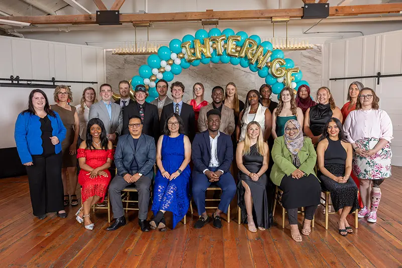 Group photo of about two dozen young professionals dressed formally, posed under a teal and gold "Intern GF" balloon arch.