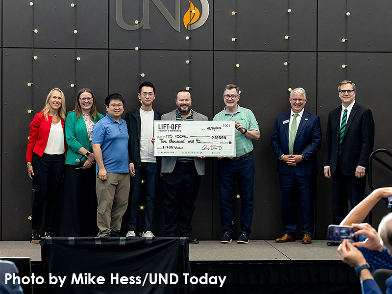 Eight people on a stage at UND holding an oversized "Lift Off" check for $10,000 awarded to ND Vocal, with UND signage behind them.