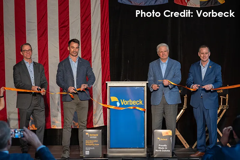 Four men in business attire stand on a stage during the Vorbeck ribbon-cutting ceremony, holding an orange ribbon stretched in front of a podium displaying the blue and yellow Vorbeck logo, with a large American flag hanging as a backdrop and product display signs reading "Next Generation PFAS-Free Firefighting Foam" and "Proudly Made in North Dakota" flanking the podium.