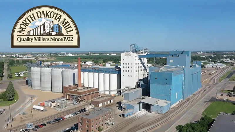 Aerial view of the North Dakota Mill and Elevator facility in Grand Forks, showing large grain silos, blue industrial buildings, and rail lines, with the North Dakota Mill logo overlaid in the upper left corner.