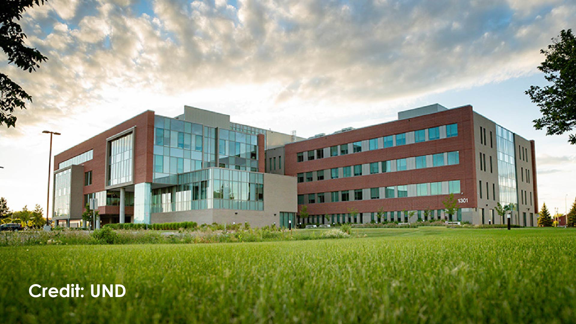 A modern red-brick and glass University of North Dakota biomedical research building rises against a partly cloudy sky at golden hour, with extensive multi-story glass facades, a manicured green lawn in the foreground, and trees framing the edges of the image.