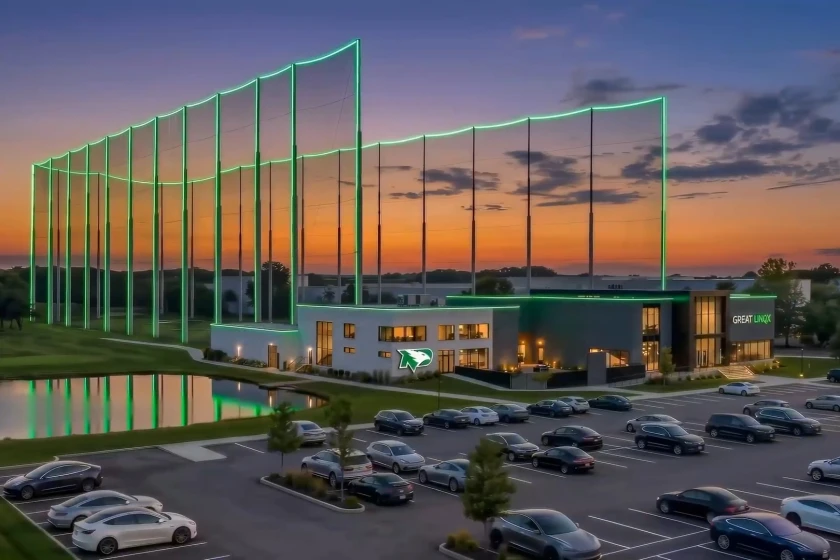 Architectural rendering of a golf entertainment venue at dusk, with illuminated green netting poles, a lit modern building, and a parking lot.